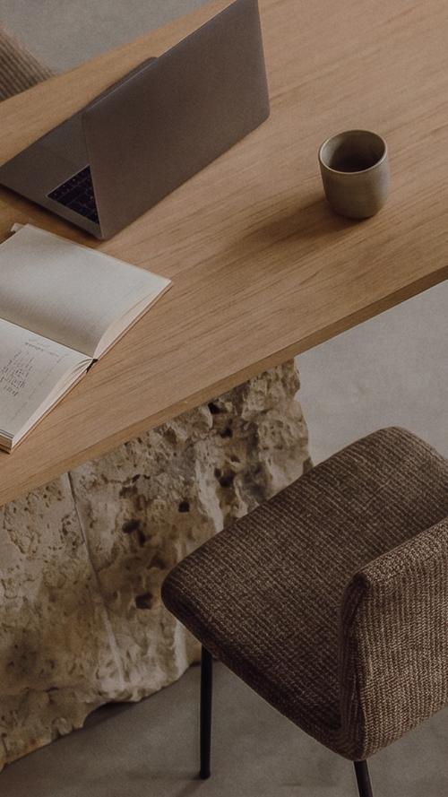 An overhead view of a wooden desk with an open laptop, a handwritten notebook, a ceramic cup, and a woven chair tucked underneath.