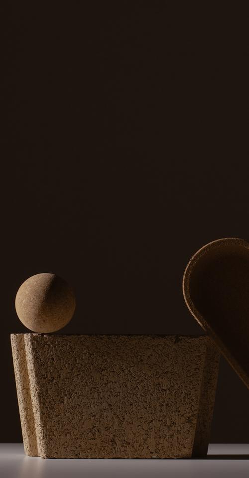 A small sphere balanced on the rim of a tapered, cork-textured vessel beside a leaning oval tray, lit against a dark brown backdrop