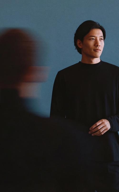 Image of a man standing against a blue wall with blurred people in the foreground.