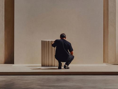 A man in dark navy crouching beside a tall sculptural plaster column in a sunlit gallery space with warm cream walls.