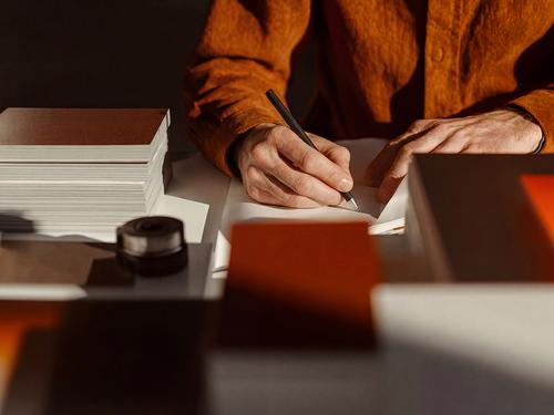 A close-up of hands in a rust-orange shirt writing in a small bound notebook on a desk strewn with stacked books and craft materials.
