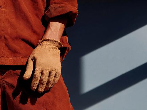 A close-up of a hand wearing a tan work glove resting against the hip of a person in rust-orange workwear, lit by a sharp diagonal shaft of sunlight.