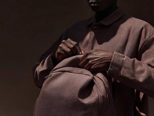 A person in a brown overshirt holding a folded fabric bag close to their chest, photographed in low warm light against a deep brown background.