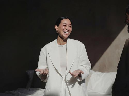 A woman in an oversized white blazer laughing with her hands gestured open, photographed against a warmly lit beige and dark backdrop.
