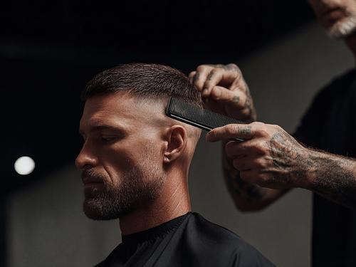 Gros plan d’un homme élégant barbu avec une coupe de cheveux dégradée qui reçoit un dernier coup de peigne de la part d’un barbier aux mains tatouées.