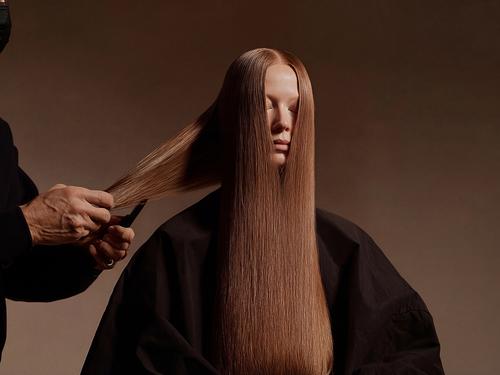 Les mains d’un coiffeur coupant les cheveux longs, raides et auburn d’une femme, qui est assise, drapée, les yeux fermés.