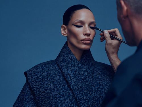 Un maquilleur appliquant un eyeliner foncé sur la paupière d’une mannequin, qui a un maquillage fort et dramatique et porte une veste bleue texturée.
