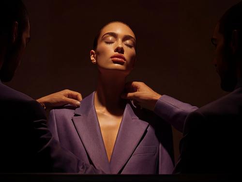Une femme aux cheveux plaqués en arrière et vêtue d’un blazer violet se tenant entre les mains de deux hommes qui ajustent le col de sa veste.