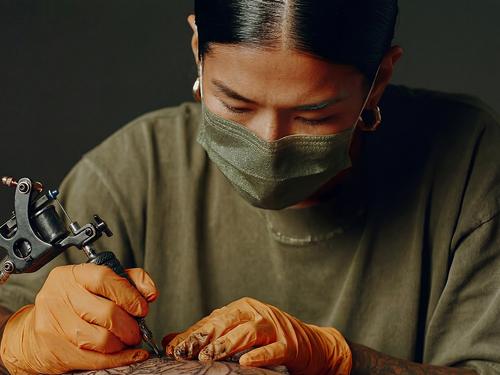 Une tatoueuse portant un masque vert et des gants orange travaillant intensément avec une machine à tatouer sur la peau d’un client.