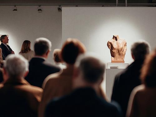 A group of people viewing a spotlighted terracotta torso sculpture in a bright, modern art gallery or museum.