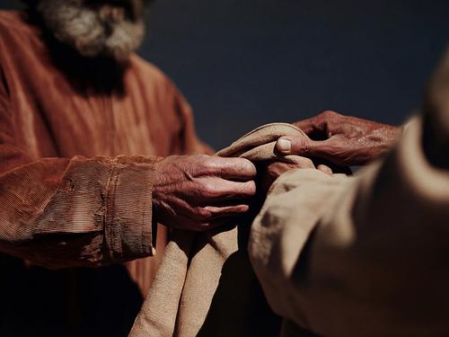A close-up shot of two people's hands, one with rougher skin, gently folding or handling a piece of rough, light brown cloth.