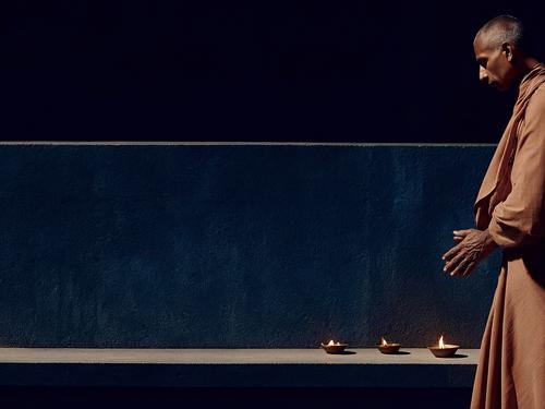 A man in simple brown robes performing a ritual with three lit oil lamps on a stone bench against a dark blue background.