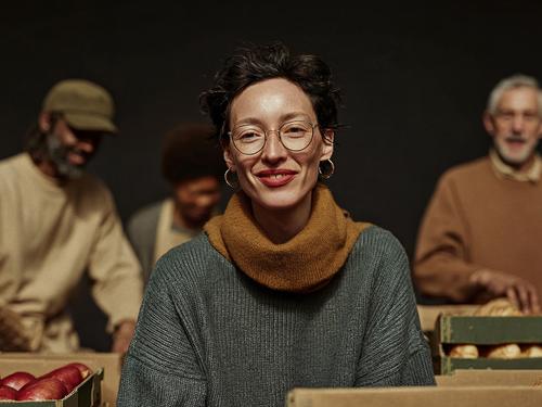 A smiling woman wearing glasses and a scarf, looking at the camera, with blurred diverse people working in the background, surrounded by boxes of food.