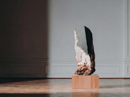 何もない白い部屋の木のブロックの上に展示されている、表面が青灰色と黄褐色の、大きくて高さのある長方形の石の彫刻。