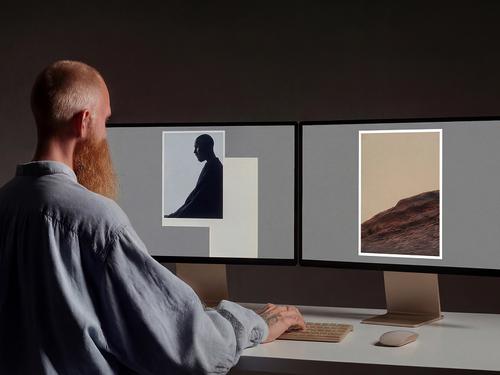 A bearded man working at two computer monitors displaying graphic design layouts with abstract shapes and photography.