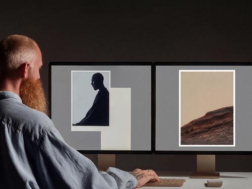 추상적인 도형과 사진의 그래픽 디자인 레이아웃이 담긴 두 대의 컴퓨터 모니터 앞에서 작업 중인 수염을 기른 남성