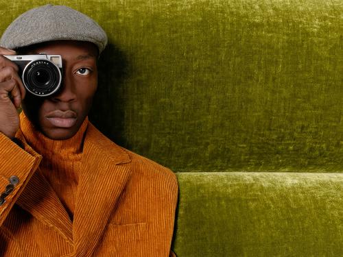 화사한 녹색 질감의 배경 앞에서 오렌지색 코듀로이 재킷과 플랫 캡을 착용한 채 카메라를 눈에 대고 있는 세련된 흑인 남성의 클로즈업.