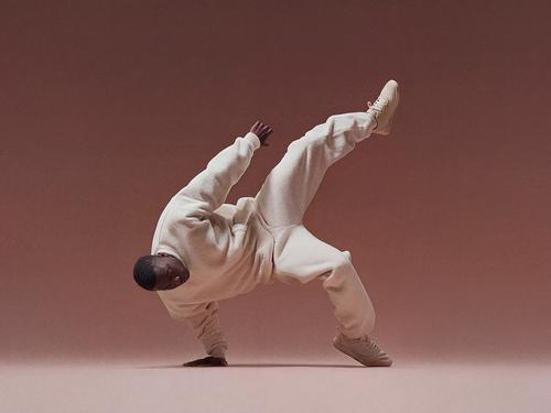 미색 트레이닝복을 입은 남성 댄서가 적갈색 배경에서 한 손으로 균형을 잡으며 활기찬 브레이크댄스 동작을 선보이고 있는 모습