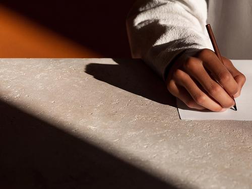 돌 책상 위에서 연필로 종이에 글씨를 쓰는 사람의 손을 극적인 각도의 조명 아래 클로즈업으로 촬영한 장면