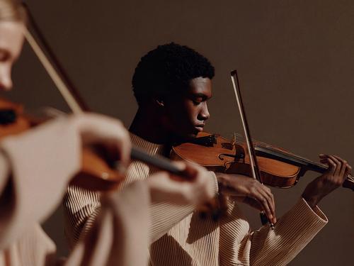 젊은 흑인 남성이 바이올린 연주에 집중하고 있고, 전경에는 흐릿하게 보이는 다른 음악가가 있으며, 두 사람 모두 밝은 색 주름 셔츠를 입고 있는 모습
