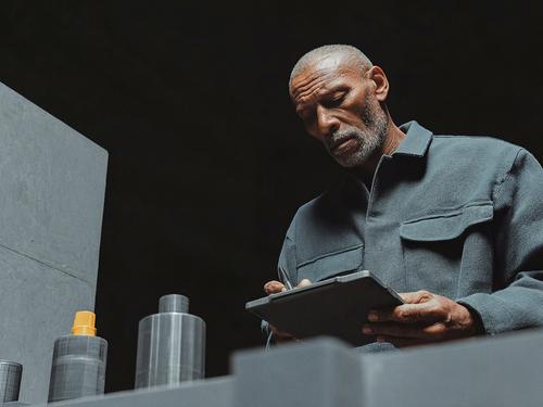 수염을 기르고 삭발한 머리를 한 나이든 남성이 짙은 파란색 작업복을 입은 채 산업 현장에서 태블릿이나 클립보드에 무언가를 쓰며 아래를 내려다보고 있는 모습