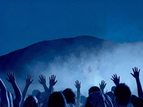 야외 콘서트 또는 쇼에서 어두운 언덕을 배경으로 푸른 빛과 안개에 둘러싸인 무대를 향해 손을 들고 있는 관중들