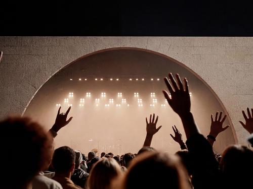 Eine große Menschenmenge auf einem Festival mit erhobenen Händen, die als Silhouetten vor einer nebligen Bühne mit hellem weißem Licht unter einem Betonbogen zu sehen sind.