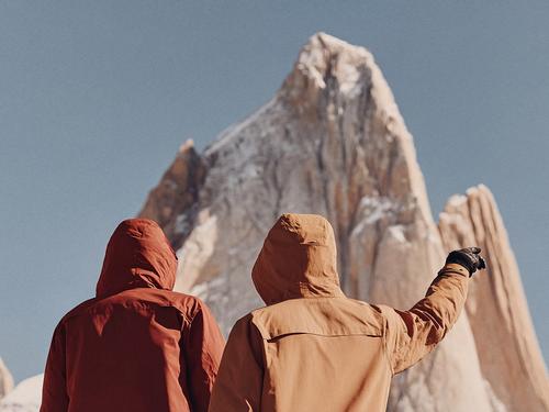 후드가 달린 재킷을 입은 두 명의 등산객이 맑고 푸른 하늘을 배경으로 눈 덮인 거친 산봉우리를 올려다보고 있는 모습