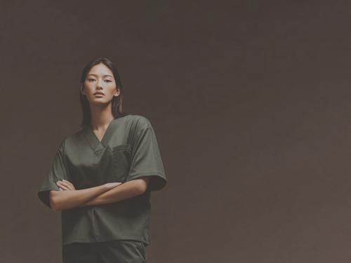 짙은 녹색의 의료진 유니폼을 입은 젊은 여성이 팔짱을 낀 채 갈색 배경에서 자신감 있게 카메라를 바라보고 서 있는 모습