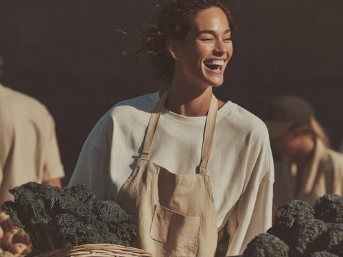 앞치마를 입고 짙은 녹색 브로콜리가 담긴 바구니 뒤에 서 있는 행복한 여성의 모습으로, 신선하고 건강한 식품 이미지를 연상시키는 밝은 분위기의 사진