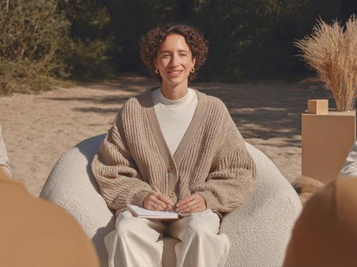 미소 짓는 치료사 또는 상담사가 야외에서 편안한 둥근 의자에 앉아 노트를 든 채 그룹 세션 참가자들에게 말을 건네는 모습