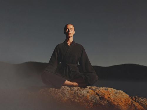 안개가 자욱한 어두운 풍경을 배경으로 이끼가 낀 커다란 바위 위에서 짙은 색 가운을 입고 다리를 꼰 채 명상하는 여성