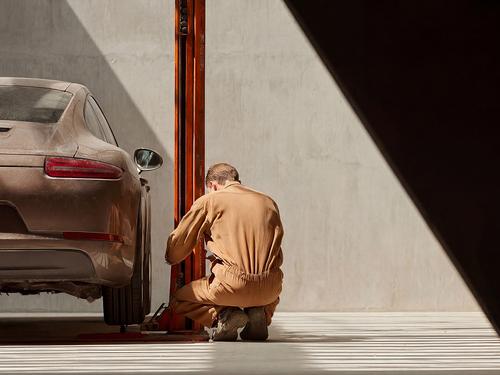 햇살이 비치는 차고에서 베이지색 작업복을 입은 정비사가 들어 올려진 갈색 스포츠카의 바퀴 부분을 수리하는 모습