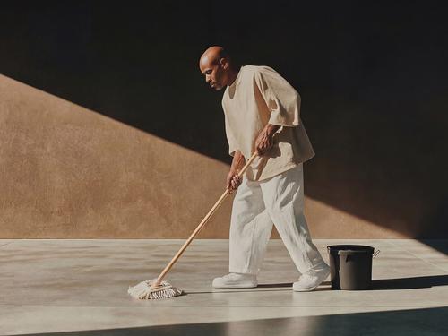 Un homme chauve portant des vêtements clairs passe la serpillière sur un grand sol intérieur à côté d’un seau noir, avec un fort contraste d’ombre et de lumière.