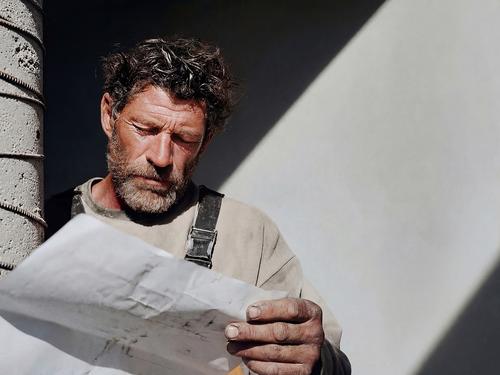 Un prestataire concentré, barbu et portant une salopette de travail, regardant un grand nombre de plans ou de documents sous une forte lumière du soleil.