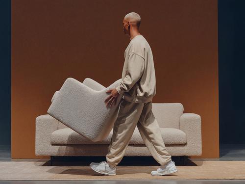 Une personne aux cheveux blonds courts portant un fauteuil blanc texturé devant un canapé blanc assorti sur un fond orange.