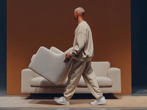 Une personne aux cheveux blonds courts portant un fauteuil blanc texturé devant un canapé blanc assorti sur un fond orange.