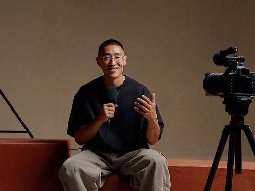 Uomo sorridente con gli occhiali siede di fronte a una videocamera professionale montata su un treppiede, mentre impugna un microfono nell'atto di registrare un video.