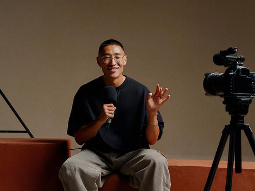 Ein lächelnder Mann mit Brille sitzt vor einer professionellen Kamera auf einem Stativ und hält ein Mikrofon, als würde er ein Video aufnehmen.