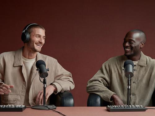 Zwei Männer sitzen an einem Tisch mit professionellen Mikrofonen und Audiomischpulten, lachen und unterhalten sich, während einer von ihnen Over-Ear-Kopfhörer trägt.