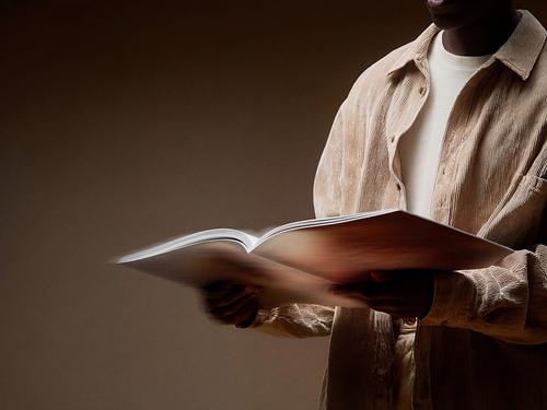 Nahaufnahme einer Person von der Hüfte aufwärts, die ein sandfarbenes Cordhemd trägt und ein großes offenes Buch oder Portfolio in den Händen hält und darin blättert.