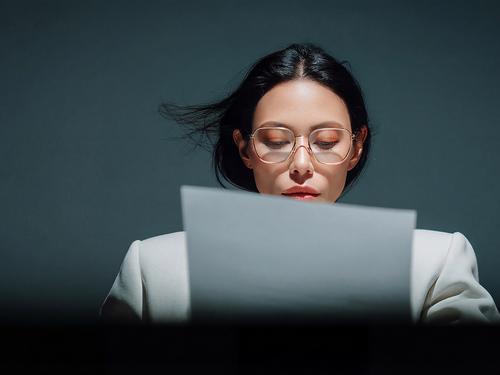 Eine Frau mit dunklen Haaren und Brille, die einen weißen Blazer trägt und konzentriert auf ein weißes Blatt Papier blickt, das sie in der Hand hält.