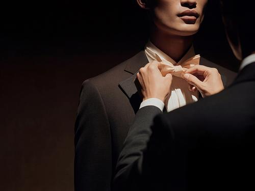 Nahaufnahme einer Person in einem dunklen Anzug, deren cremefarbene Fliege von jemand anderem in warmem, stimmungsvollem Licht zurechtgerückt wird.