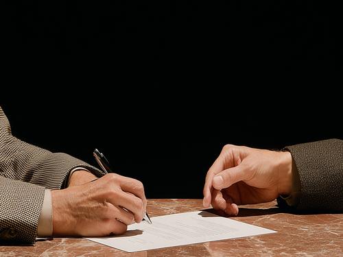 대리석 테이블 위에 놓인 두 사람의 손이 클로즈업되어 있고 한 사람이 계약서로 보이는 문서에 서명하는 모습으로, 비즈니스 상담을 연상시키는 장면