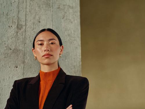 머리를 뒤로 넘긴 스타일에 주황색 터틀넥과 검은색 블레이저를 입고서 팔짱을 낀 채 콘크리트 기둥 앞에 서 있는 자신감 넘치는 여성