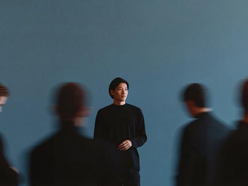 모션 블러 처리된 네 사람의 실루엣에 둘러싸인 채, 파란색 벽을 뒤로 하고 중앙에 서 있는 검은 스웨터를 입은 남성에 포커스가 맞춰져 있는 모습