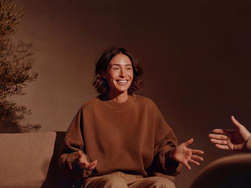 갈색 스웨터를 입은 여성 코치 또는 치료사가 소파에 앉아 미소를 지으며 상담 중 고객을 향해 두 손을 벌려 제스처를 취하는 모습