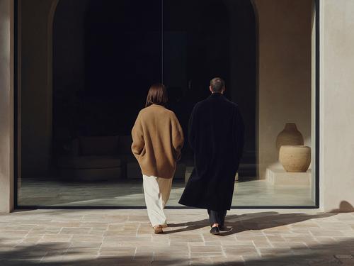 한 사람은 베이지색 스웨터를, 다른 한 사람은 검은색 코트를 입고 대형 창문이 있는 현대적이고 고급스러워 보이는 집의 출입구를 지나 걷는 두 사람의 뒷모습