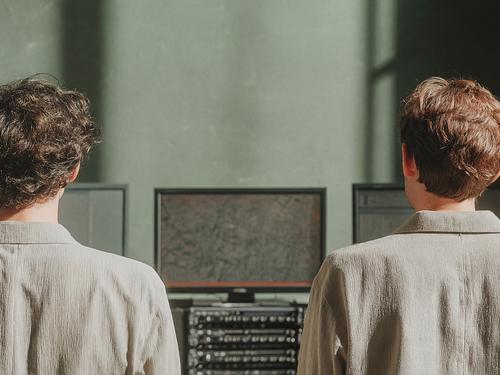 밝은 색 버튼업 셔츠를 입고 녹음실이나 기술 스튜디오에서 컴퓨터 모니터와 오디오 장비 랙을 보고 있는 두 사람의 뒷모습