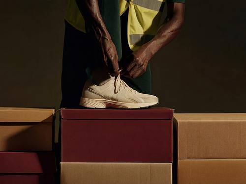 Mãos de uma pessoa amarram os cadarços de um tênis esportivo bege, que está sobre caixas de sapatos empilhadas nas cores marrom e bege.